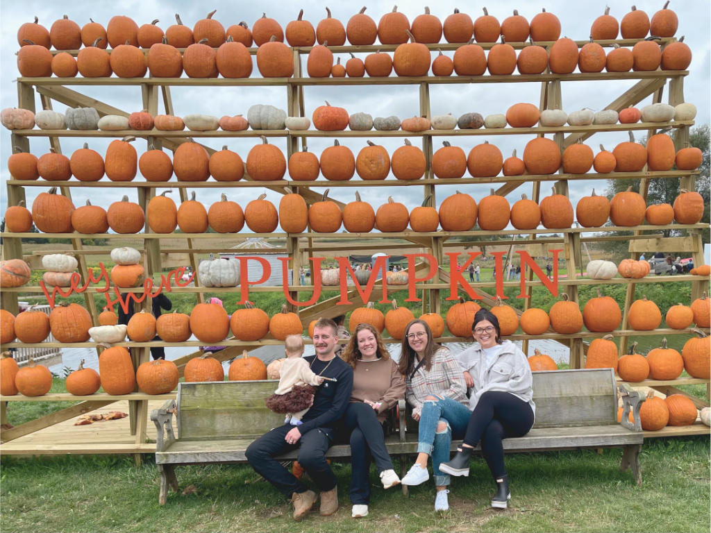 Kait with her family and friends posing in front of a pumpkin wall at an orchard